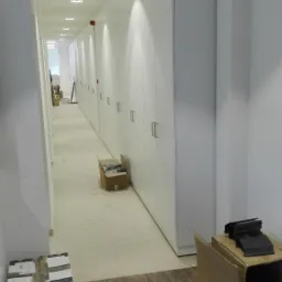 Długi korytarz z rzędem białych szaf w zabudowie, kartony z materiałami budowlanymi na podłodze z płytek imitujących drewno, widoczne ślady prac wykończeniowych.