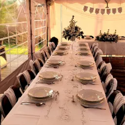 Uroczysty stół komunijny w namiocie: biały obrus z okolicznościowym nadrukiem, elegancka zastawa, dekoracje kwiatowe, balony i baner 'Komunia'.