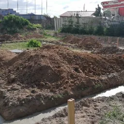 Wylane fundamenty z betonu w wykopie, z hałdami ziemi dookoła, betonomieszarka w tle.