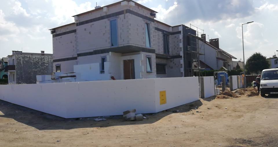 Surowy, dwupiętrowy dom z szarej cegły, w trakcie budowy, ogrodzony białymi panelami styropianowymi. Widoczne niezabezpieczone okna i fragment rusztowania.