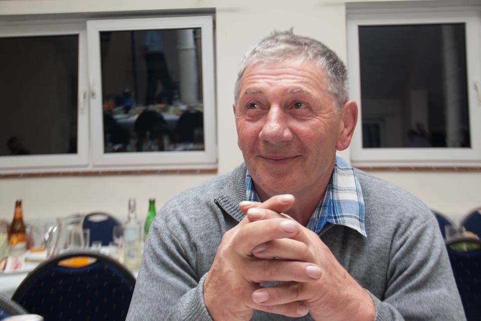 Portret uśmiechniętego mężczyzny w szarym swetrze na tle okien. Dłonie splecione, wzrok skierowany w górę. Na stole butelki i naczynia.