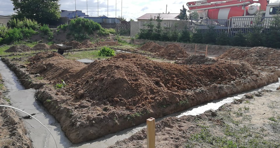 Wylane fundamenty z betonu w wykopie, z hałdami ziemi dookoła, betonomieszarka w tle.