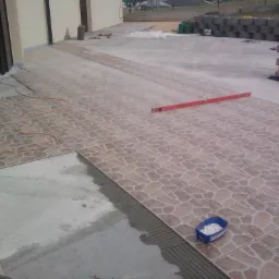 Układanie płytek ceramicznych na tarasie, widoczny poziomica, pojemnik z krzyżykami dystansowymi i częściowo ułożona podłoga z wzorem kamienia.