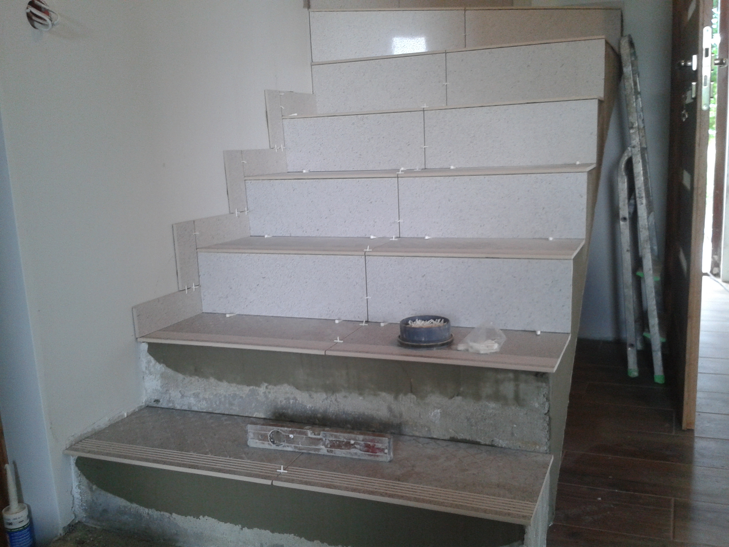 Schody w trakcie układania płytek, widoczne poziomica, pojemnik z krzyżykami dystansowymi, drabina oparta o ścianę.