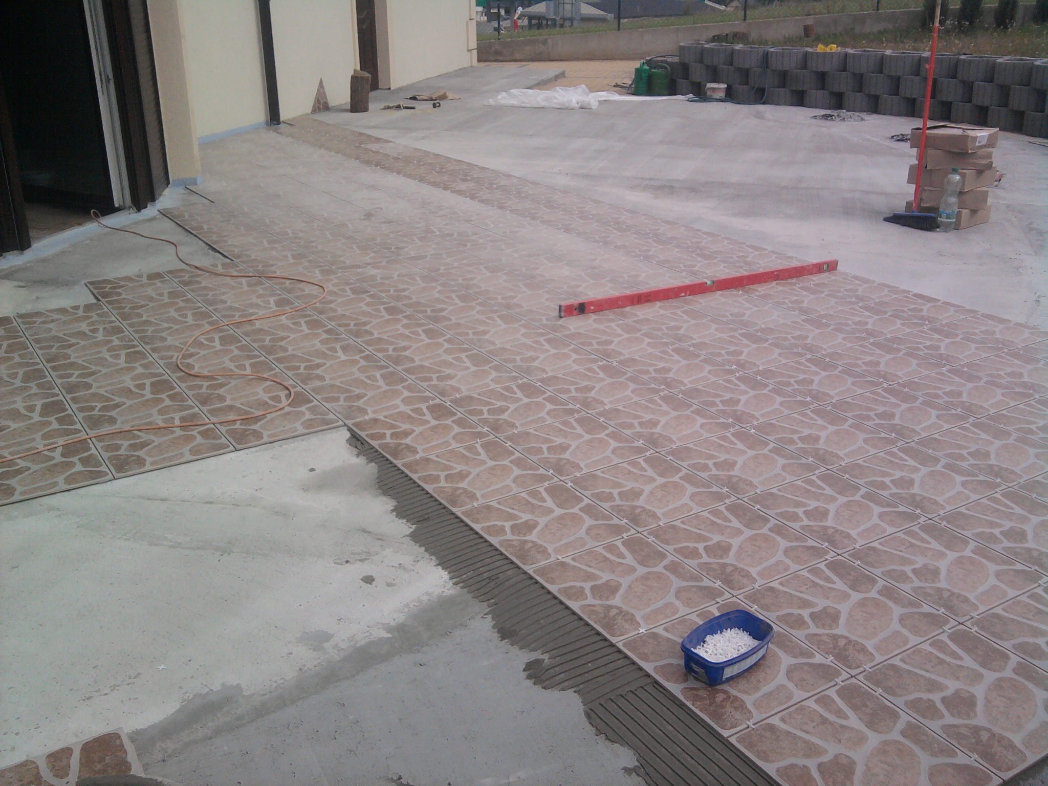 Układanie płytek ceramicznych na tarasie, widoczny poziomica, pojemnik z krzyżykami dystansowymi i częściowo ułożona podłoga z wzorem kamienia.