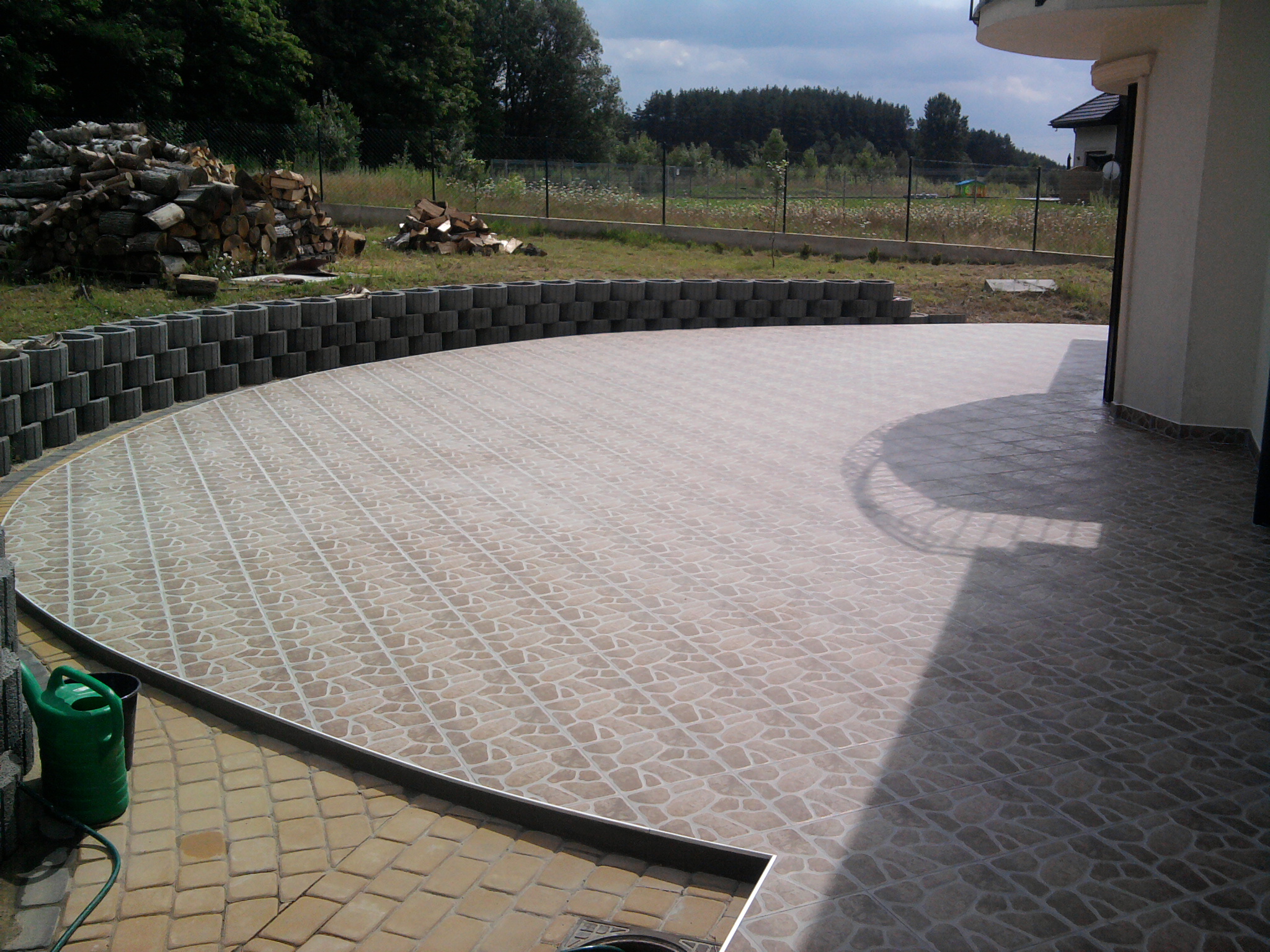 Wyłożony płytkami taras przylegający do budynku, z widocznym stosem drewna i zielonymi roślinami w tle.