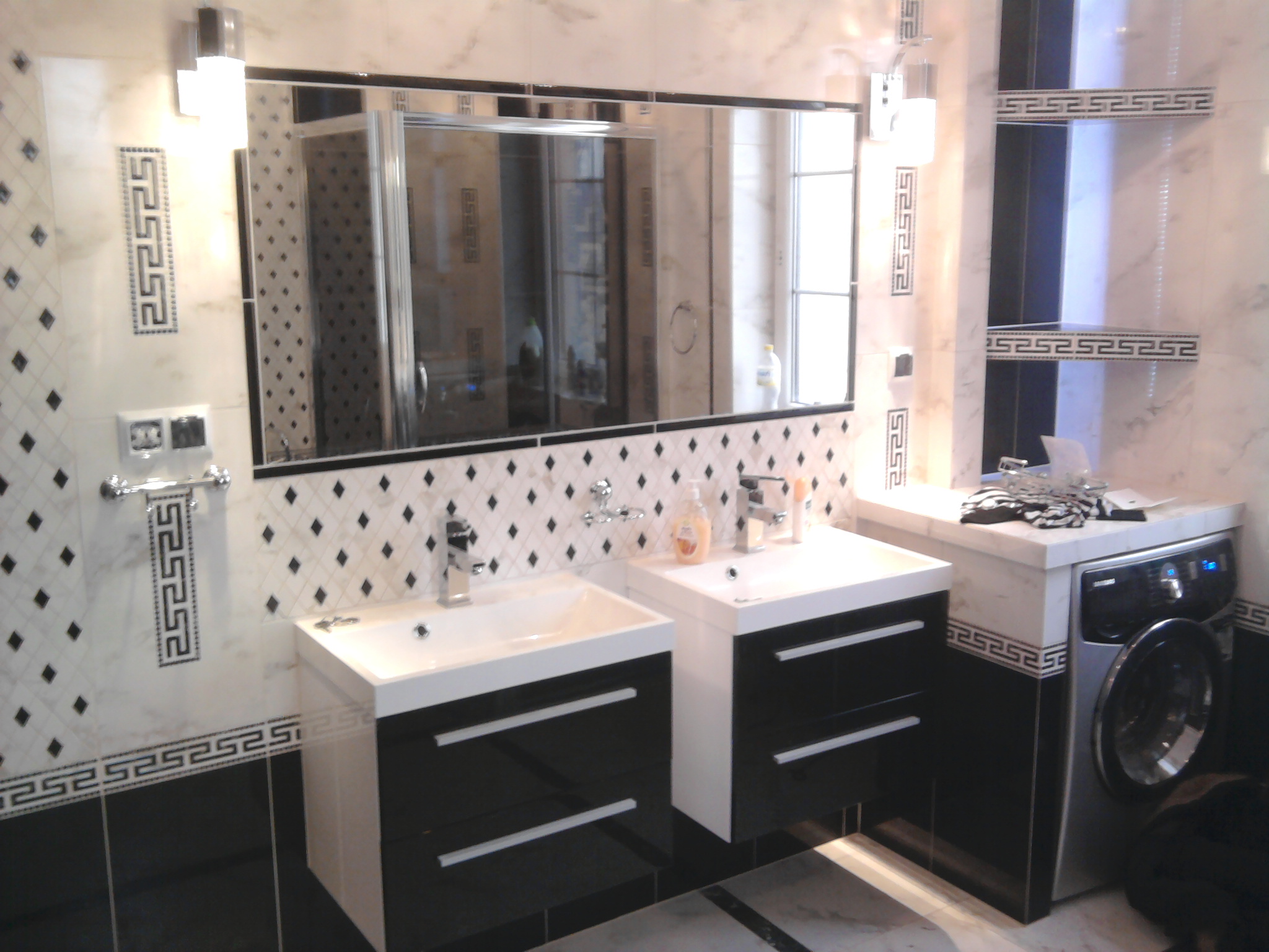 Nowoczesna łazienka w czarno-białej kolorystyce z dwoma umywalkami na szafkach z połyskiem, duże lustro z podświetleniem, pralka w zabudowie i dekoracyjne płytki z motywem greckim.