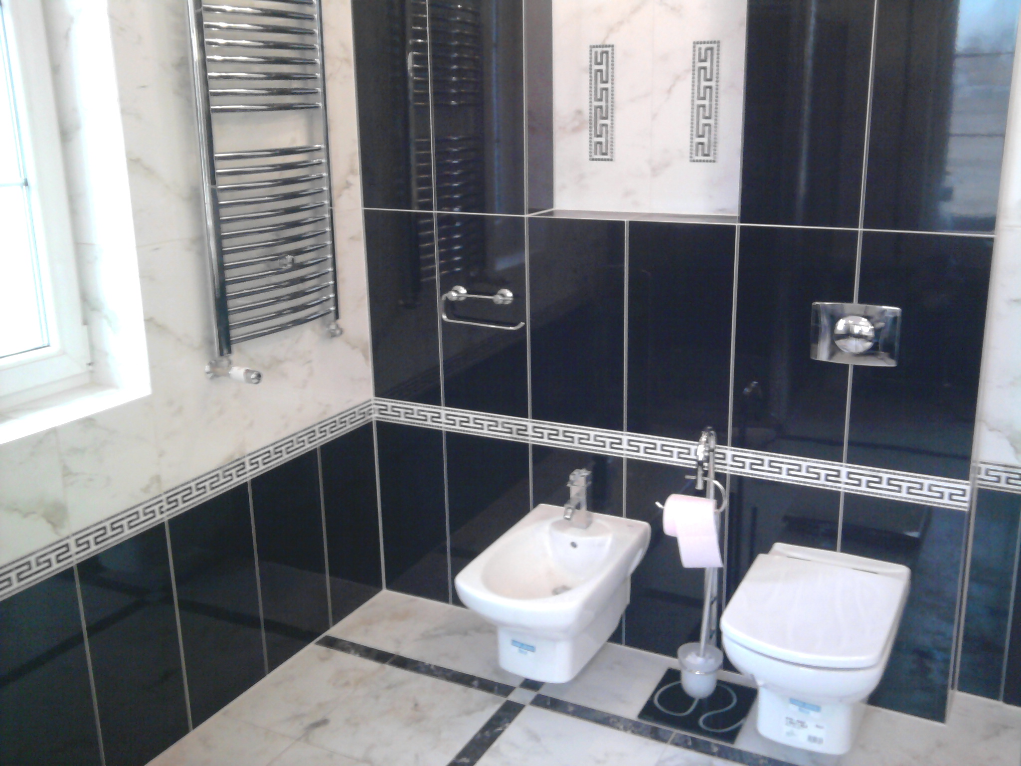 Nowoczesna łazienka z czarno-białymi płytkami, grzejnikiem drabinkowym, bidetem i toaletą podwieszaną, wykończona ozdobnym greckim motywem.