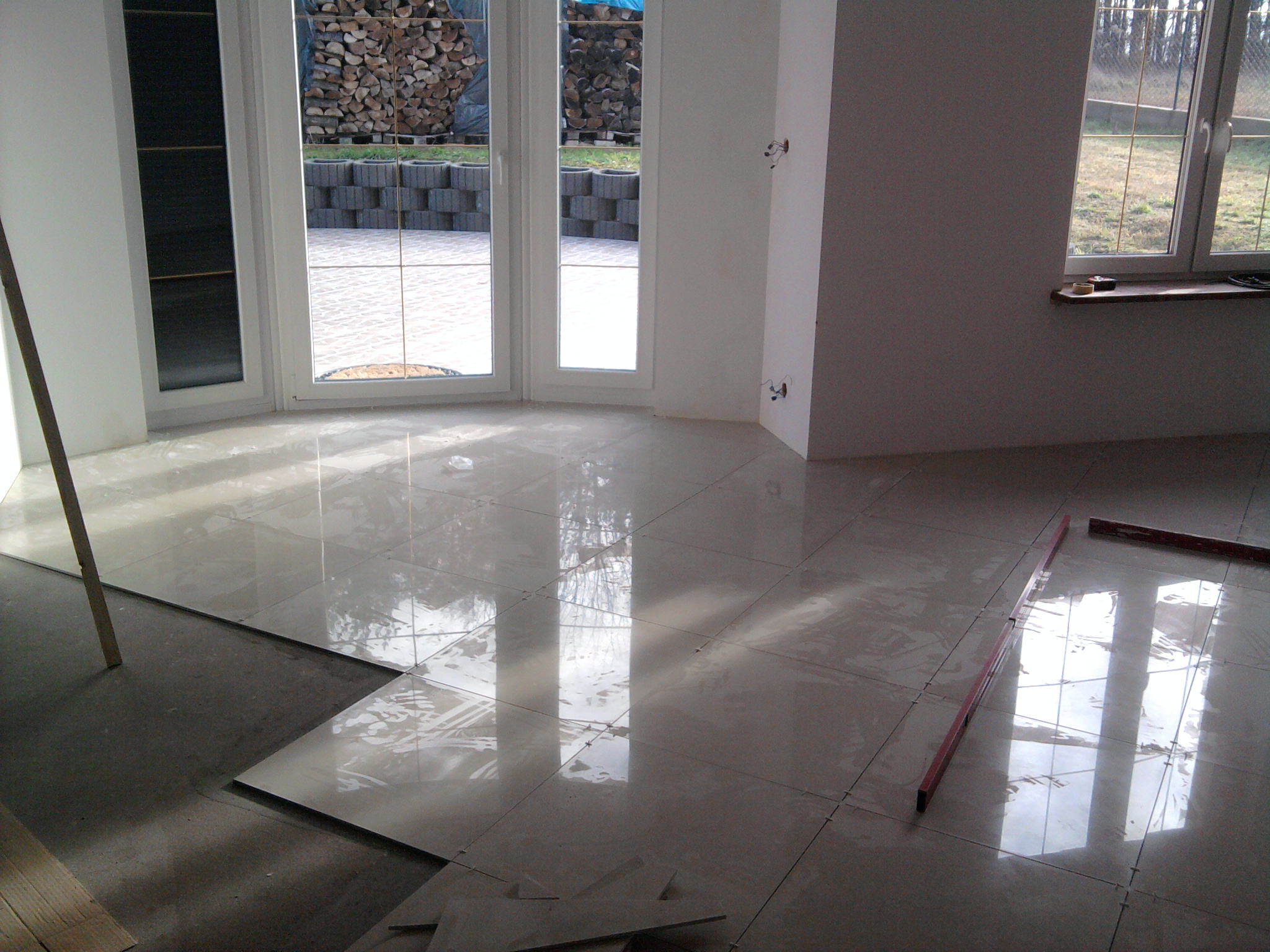 Układanie błyszczących, beżowych płytek podłogowych w nowym pomieszczeniu, widoczne poziomice i nieukończona część posadzki z drewnianymi elementami przy ścianie z oknem.