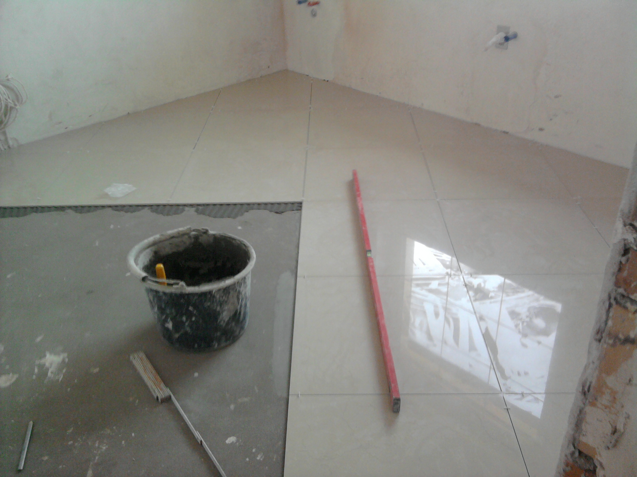 Układanie jasnych płytek podłogowych w pomieszczeniu z widocznym wiadrem z zaprawą, poziomicą i fragmentem ściany w stanie surowym