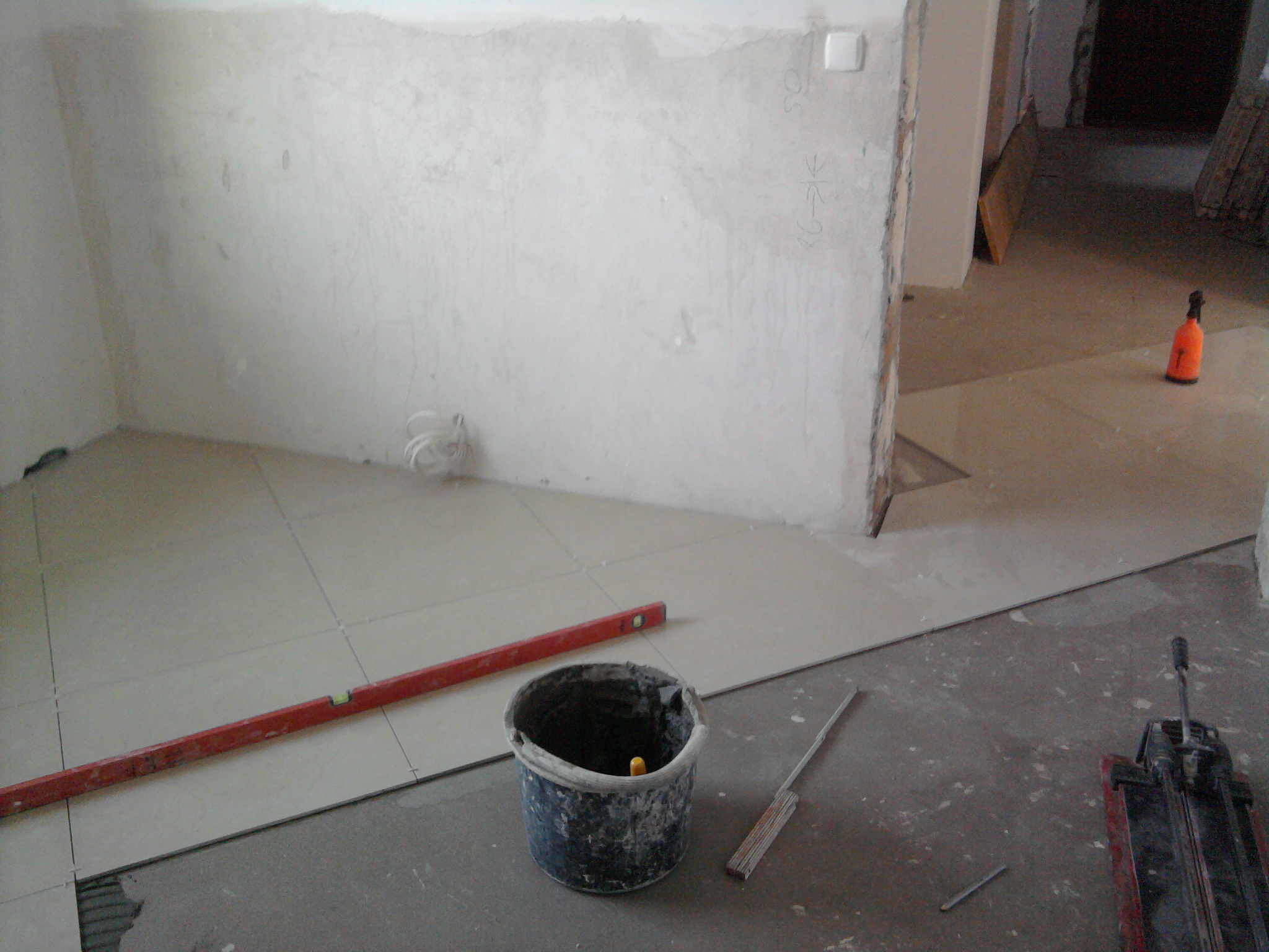 Układanie jasnych, kwadratowych płytek podłogowych w trakcie remontu, widoczny poziomica, wiadro i przecinarka do płytek, ściany w stanie surowym.