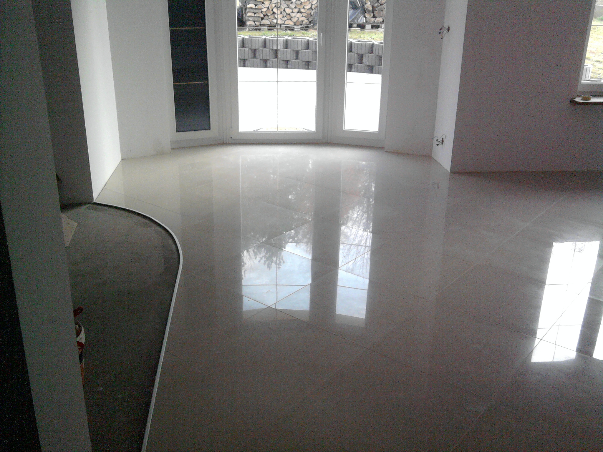 Połyskująca, beżowa podłoga z płytek w jasnym pomieszczeniu z dużym oknem; widoczny fragment niedokończonej posadzki z szarym podkładem.