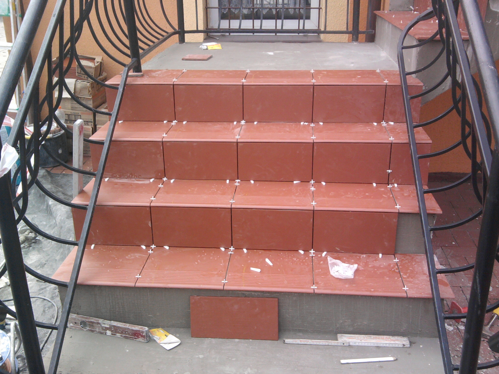 Montaż terakotowych płytek na schodach zewnętrznych z czarną, metalową balustradą, widoczne krzyżyki dystansowe i poziomica.