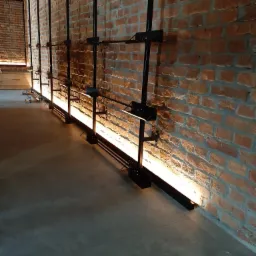Oświetlenie podłogowe LED zamontowane wzdłuż ceglanej ściany, czarne metalowe wsporniki, surowy beton na podłodze, ciepłe światło odbijające się od tekstury cegieł.