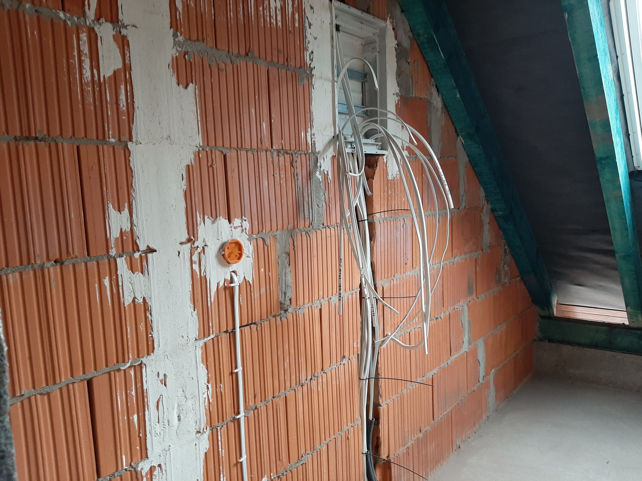 Surowa ściana z czerwonej cegły z niedokończonymi przewodami elektrycznymi wychodzącymi z puszki rozdzielczej; widoczne ślady zaprawy i gipsu.