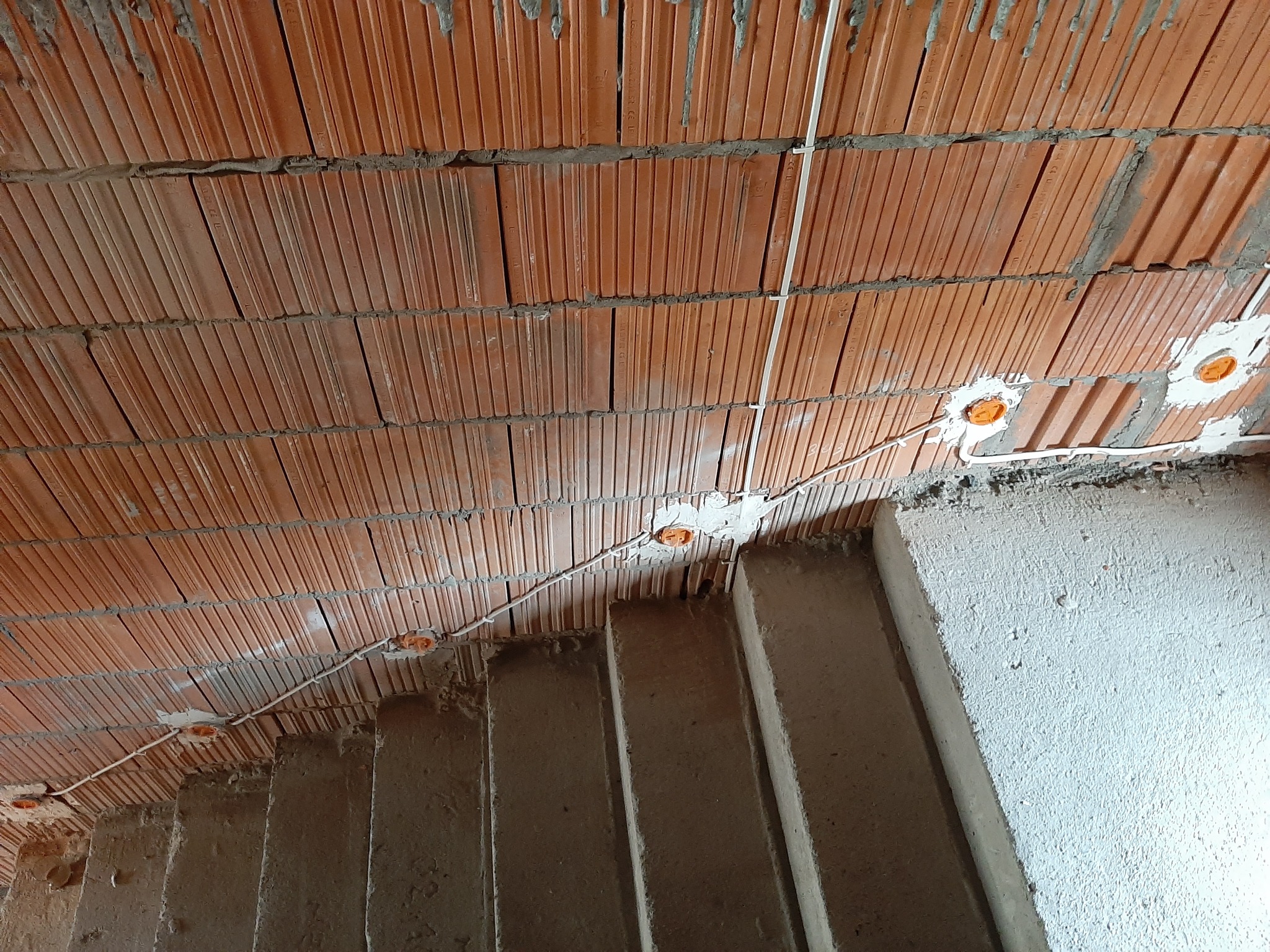 Ceglany strop z widocznymi przewodami elektrycznymi i puszkami instalacyjnymi, częściowo zasłonięty przez betonowe schody w surowym stanie.