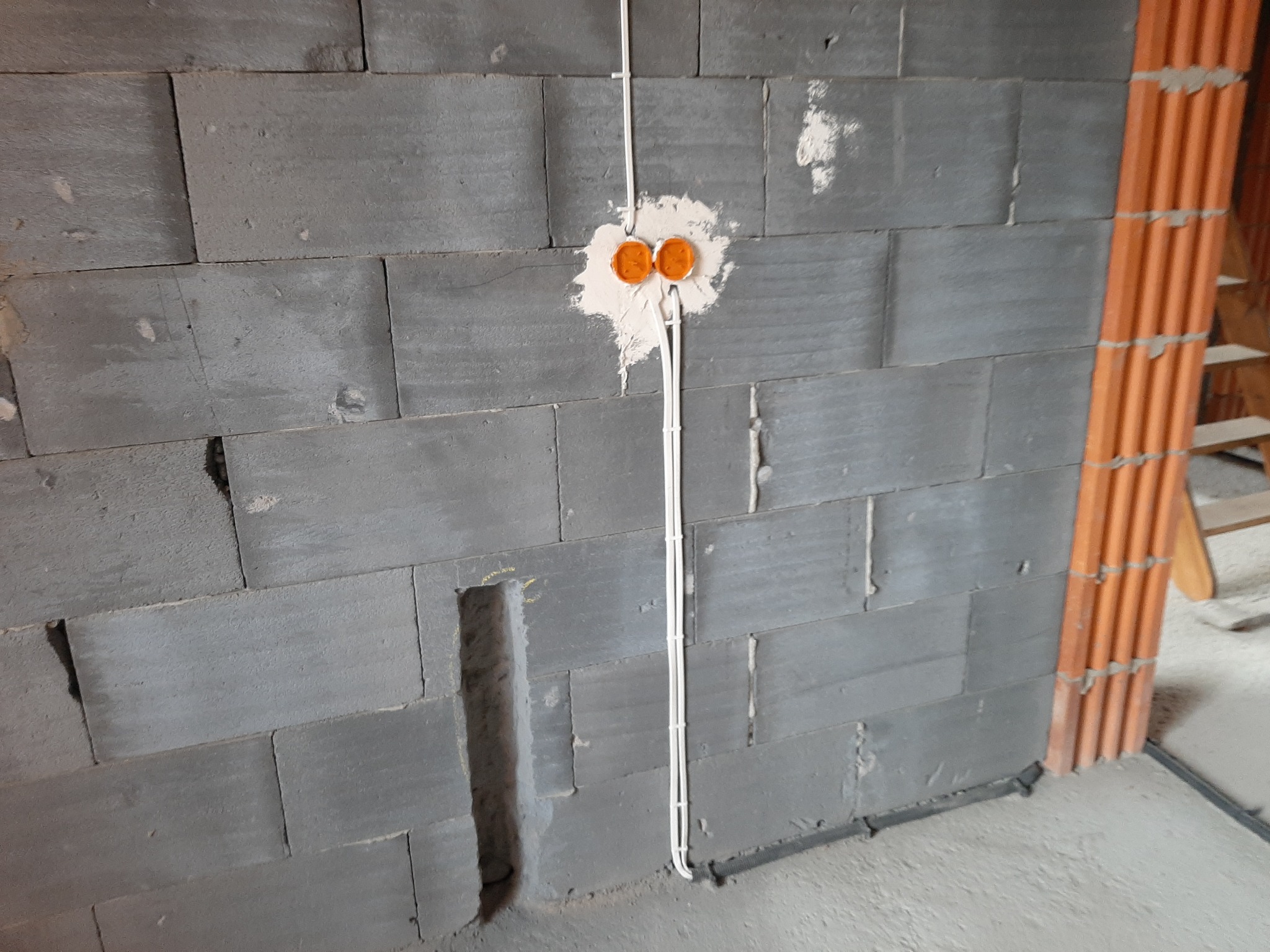 Ściana z szarego betonu komórkowego w trakcie prac instalacyjnych, widoczne puszki elektryczne w białym gipsie i pionowo poprowadzone przewody w białej izolacji.