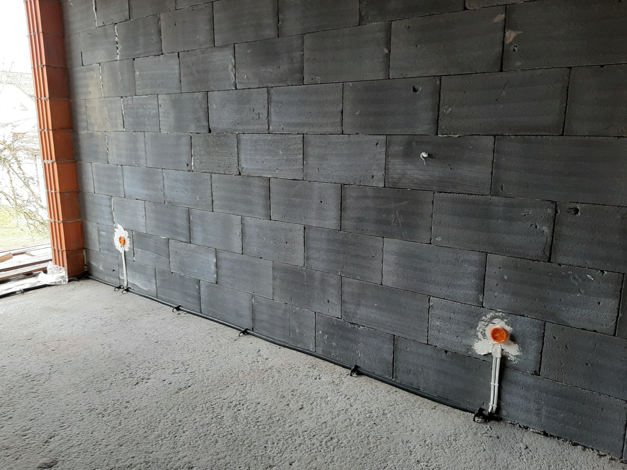 Szara ściana z betonu komórkowego w trakcie budowy z widocznymi puszkami elektrycznymi i przewodami ułożonymi wzdłuż podłogi.