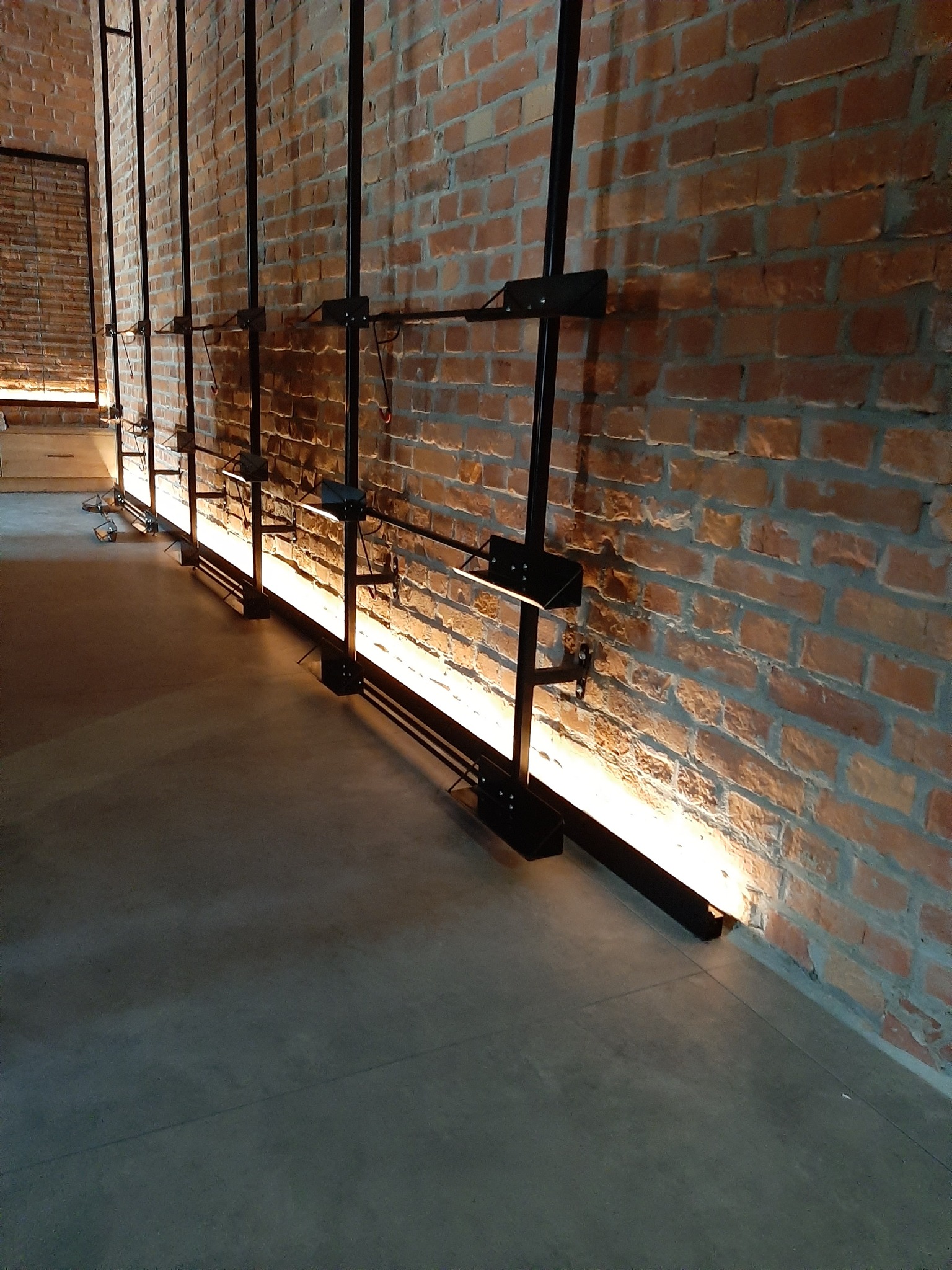 Oświetlenie podłogowe LED zamontowane wzdłuż ceglanej ściany, czarne metalowe wsporniki, surowy beton na podłodze, ciepłe światło odbijające się od tekstury cegieł.