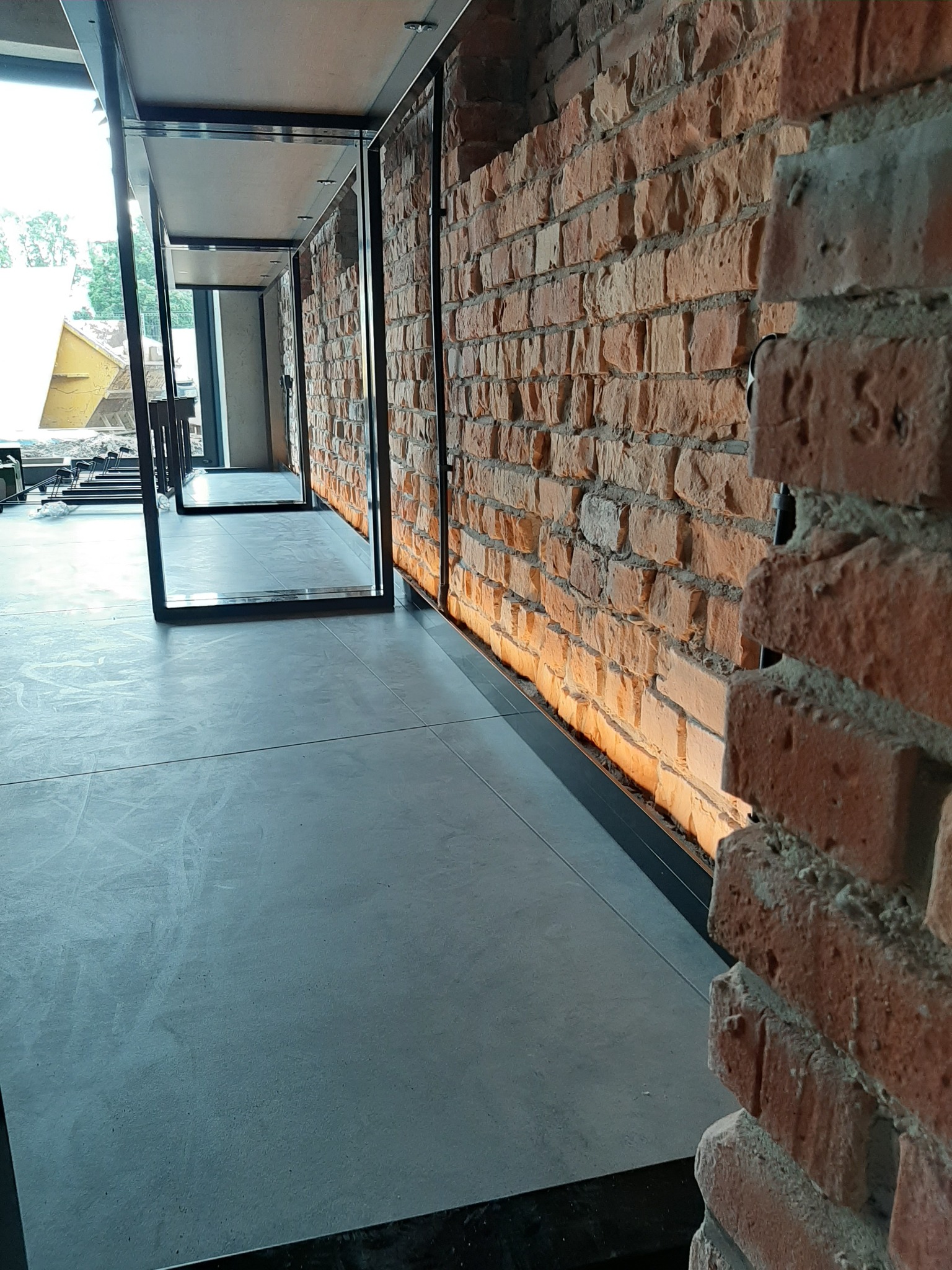 Wnętrze z surową ceglaną ścianą oświetloną od dołu, betonowa posadzka i minimalistyczne czarne ramy drzwiowe tworzące perspektywę.