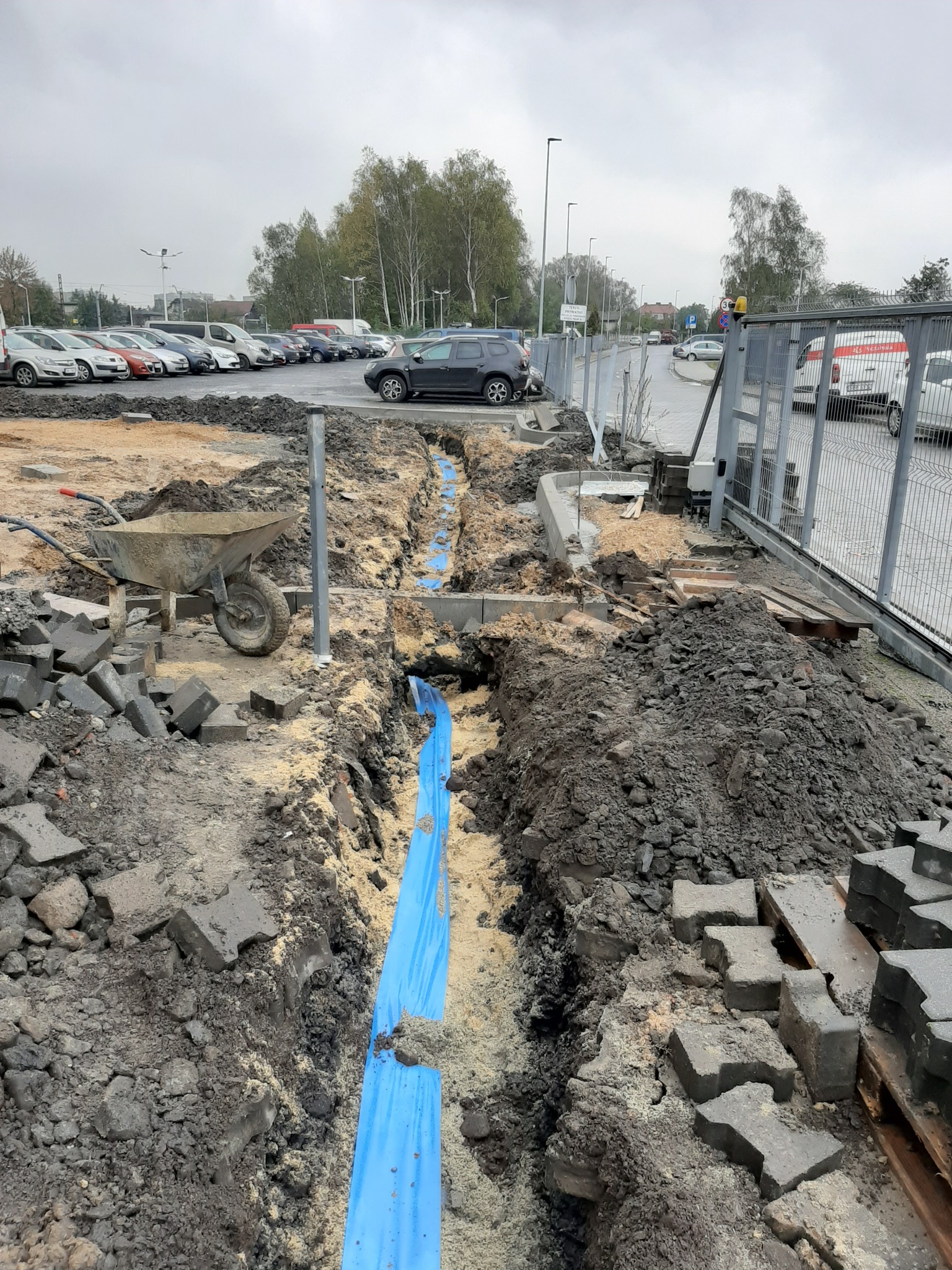 Wykop z niebieską rurą osłonową na terenie budowy parkingu, widoczne kostki brukowe, taczka i ogrodzenie z siatki.