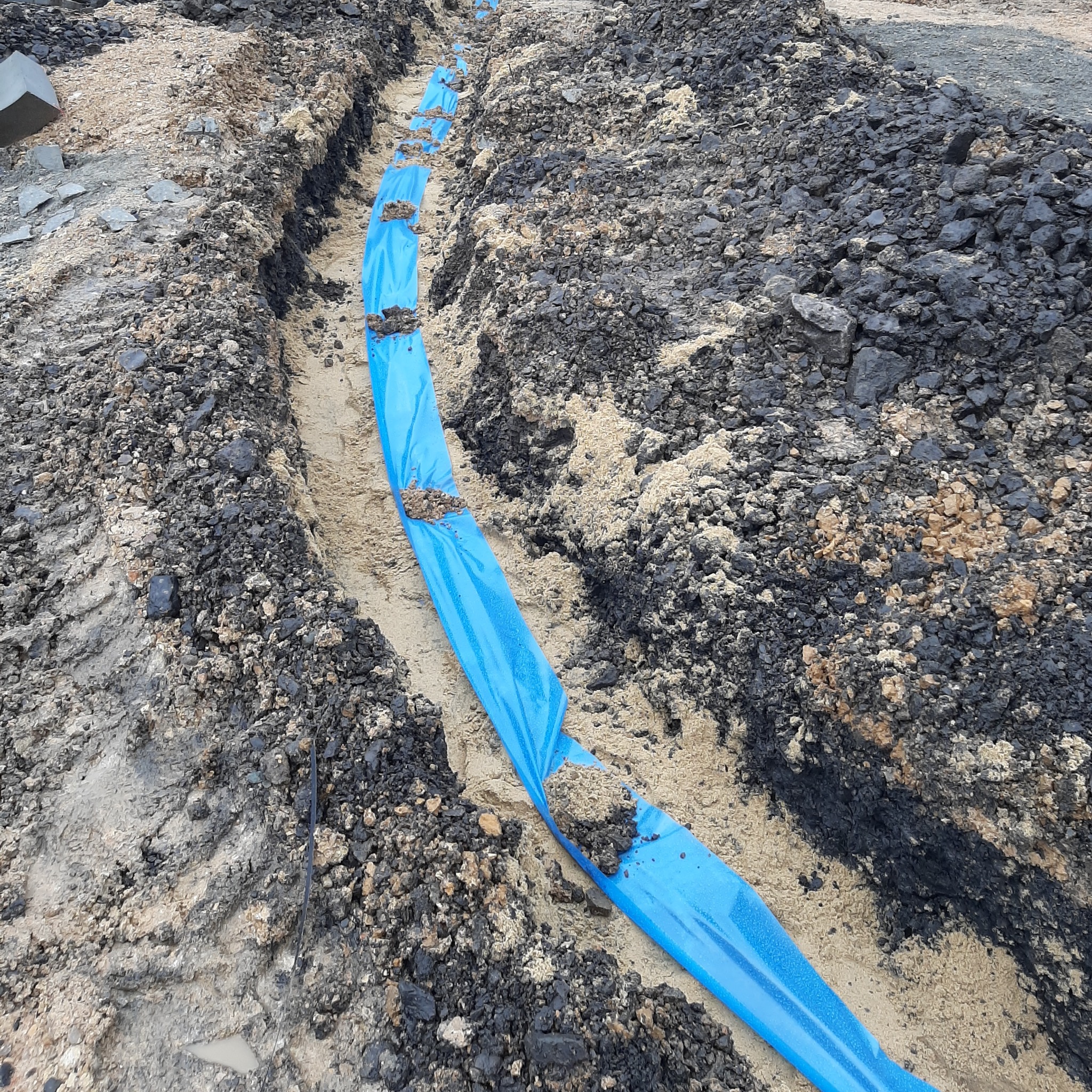 Wykop pod instalację, zabezpieczony niebieską folią, częściowo zasypany ziemią i kamieniami. Widoczne ślady opon koparki.