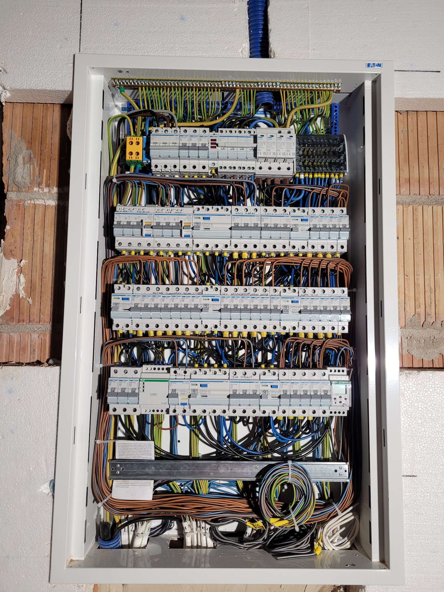 Otwarta skrzynka rozdzielcza z licznymi bezpiecznikami i uporządkowanymi przewodami w różnych kolorach, zamontowana na ścianie w surowym stanie, widoczne fragmenty cegły i styropianu