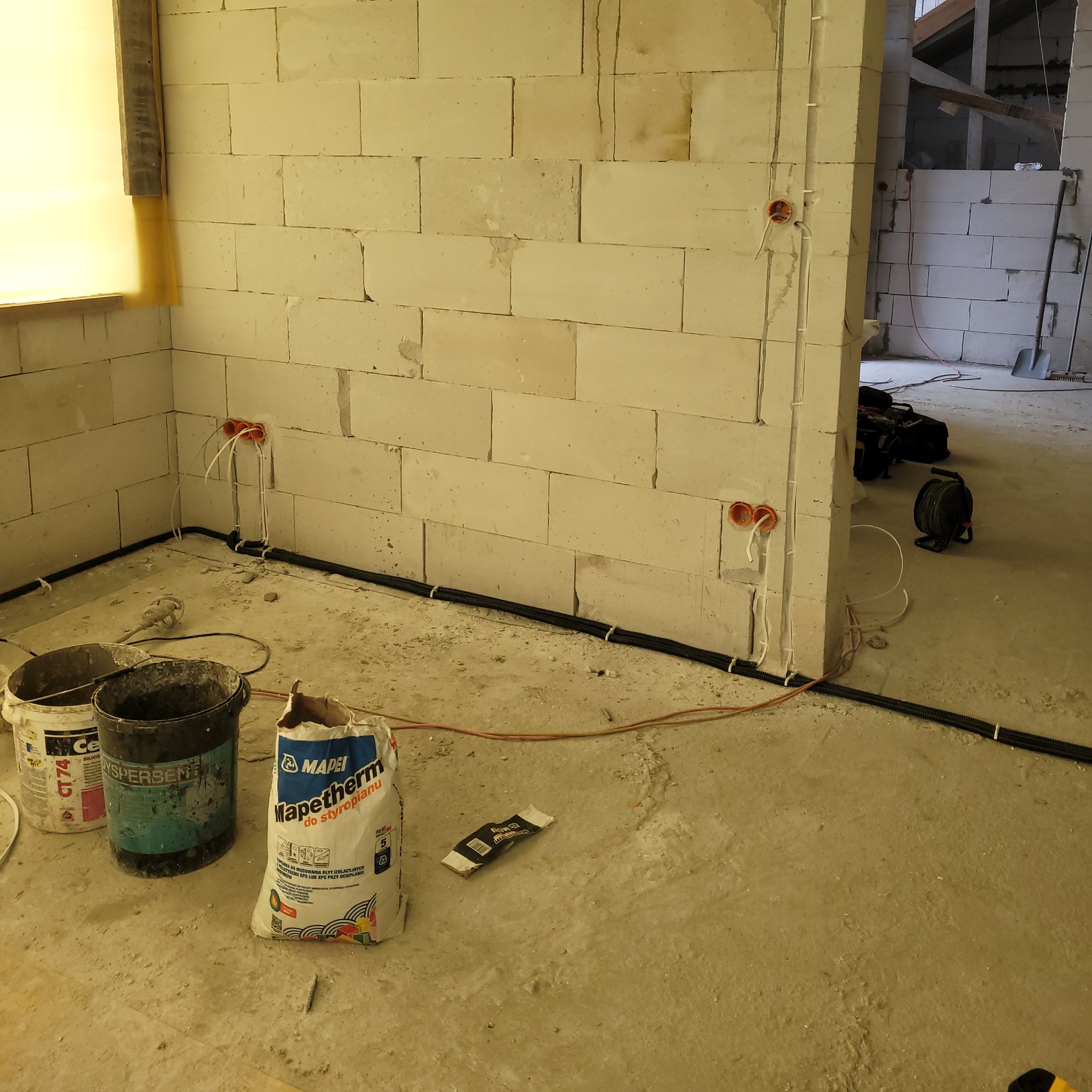 Wnętrze w trakcie remontu z widocznymi puszkami elektrycznymi w ścianie z betonu komórkowego, czarnymi rurkami karbowanymi przy podłodze, workiem zaprawy Mapei i narzędziami w tle.