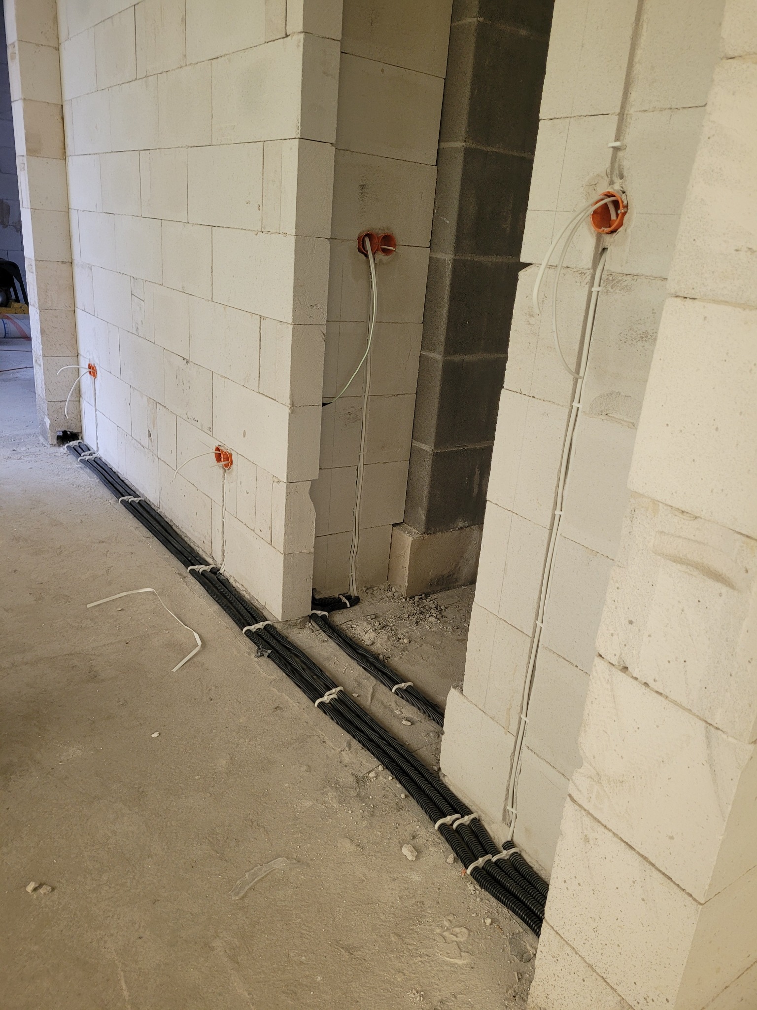 Instalacja elektryczna w surowym wnętrzu: kable w peszlach ułożone na podłodze, przewody wychodzące z pomarańczowych puszek w ścianach z białych bloczków.