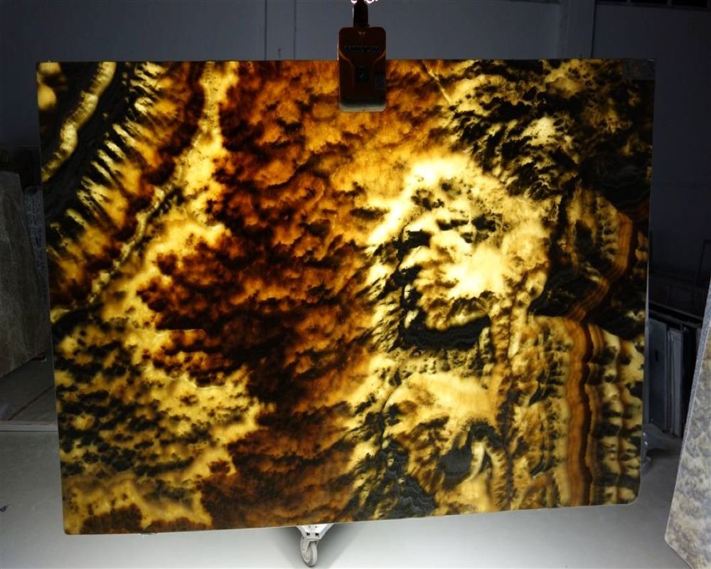 Podświetlona płyta z onyksu o nieregularnym wzorze w odcieniach brązu, czerni i bieli, zawieszona na uchwycie w magazynie kamienia.