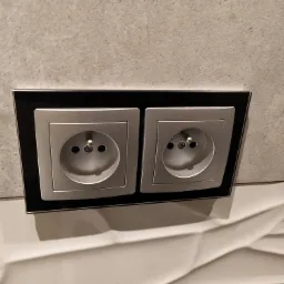 Dwa gniazdka elektryczne w srebrnej ramce z czarnym obramowaniem, zamontowane na szarej ścianie nad białą, trójwymiarową dekoracyjną płytą ścienną.