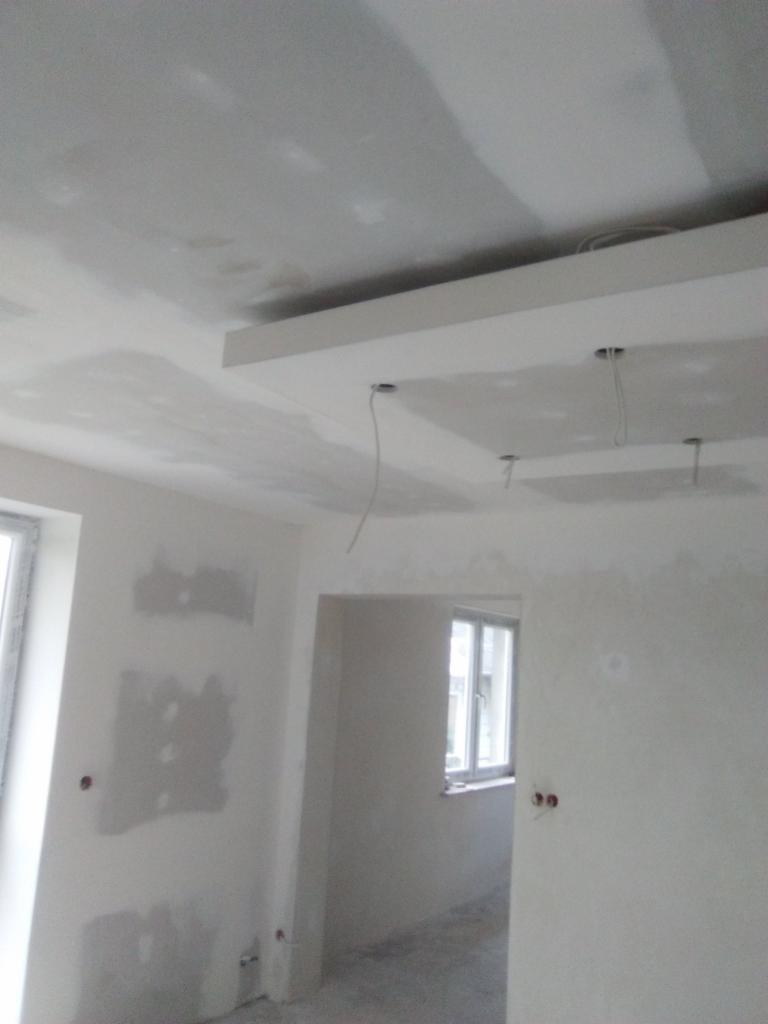 Surowe wnętrze w trakcie tynkowania ścian i sufitu z widocznymi przewodami elektrycznymi wystającymi z sufitu.