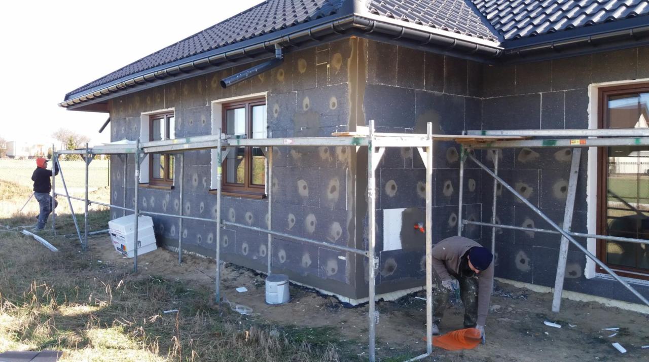 Dom jednorodzinny w trakcie ocieplania: czarne płyty izolacyjne z widocznymi punktami mocowania, fragment ściany z nałożoną warstwą zaprawy, rusztowanie, pracownicy w trakcie wykonywania prac...