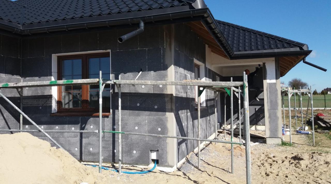 Dom jednorodzinny w trakcie prac związanych z ociepleniem elewacji, widoczne szare płyty izolacyjne na ścianach, rusztowania i piasek na pierwszym planie, fragment okna z brązową ramą.