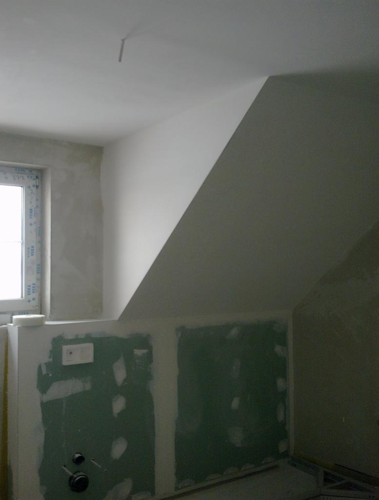 Wnętrze poddasza w trakcie prac wykończeniowych: ściany częściowo pokryte płytami gipsowo-kartonowymi, widoczne okno PCV z naklejkami producenta, sufit i ściany przygotowane do malowania.