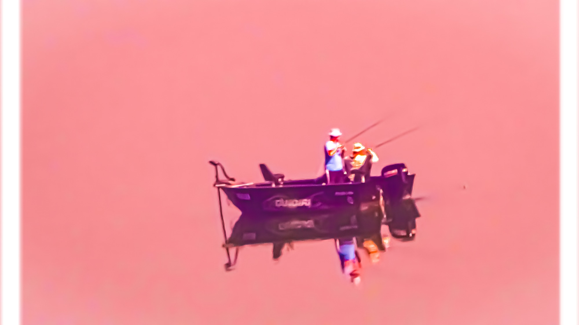 Dwóch wędkarzy łowiących ryby z ciemnej łodzi na spokojnej wodzie, widoczne wędki i odbicie łodzi w wodzie.