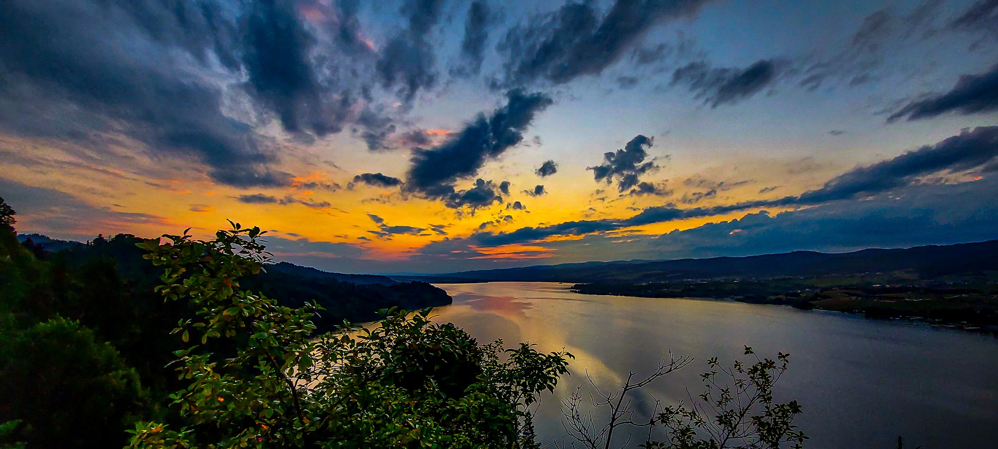 Panoramiczny widok na jezioro w blasku zachodzącego słońca, z ciemnymi chmurami i zieloną roślinnością w pierwszym planie.