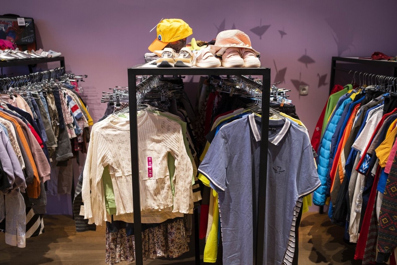 Wnętrze sklepu z odzieżą używaną: wieszaki z ubraniami, buty i czapki na półkach. Widoczne różne style i kolory ubrań oraz akcesoriów.