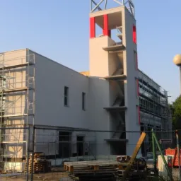 Budynek w trakcie prac elewacyjnych, widoczne rusztowania, świeżo otynkowana fasada w kolorze jasnego beżu, wieża z czerwonymi akcentami, teren budowy zabezpieczony siatką.