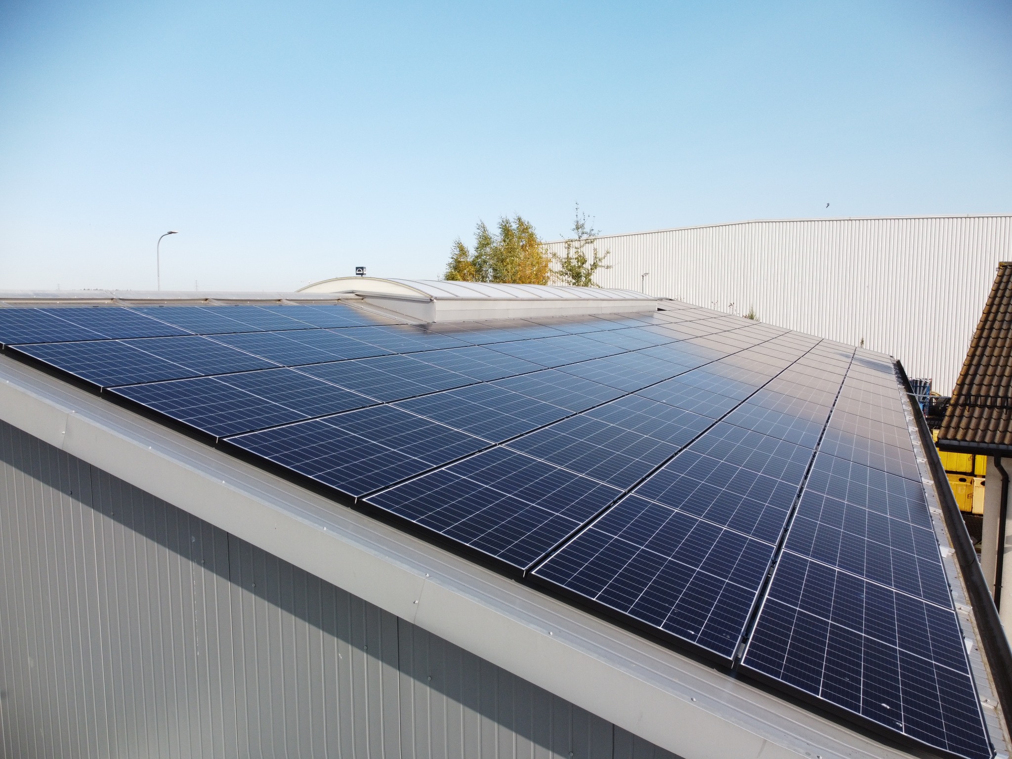 Instalacja paneli słonecznych na dachu hali przemysłowej, widok z góry, niebieskie niebo w tle.