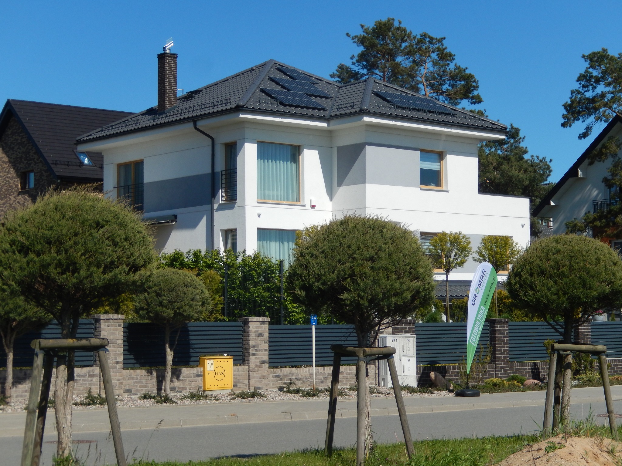 Dwukondygnacyjny dom z szarą dachówką i panelami fotowoltaicznymi na dachu, widok z ulicy, w tle zielony baner reklamowy firmy fotowoltaicznej.