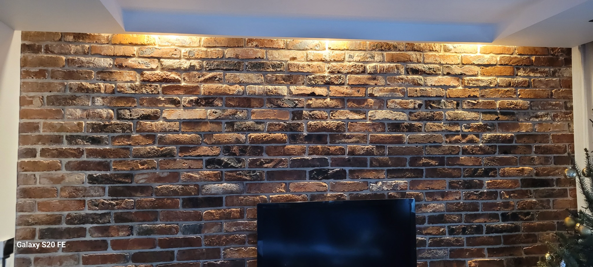Dekoracyjna ściana z cegły klinkierowej w salonie, oświetlona od góry ciepłym światłem, widoczny fragment telewizora i choinki.