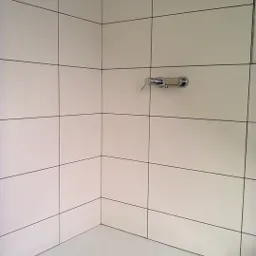 Narożnik wykafelkowanej kabiny prysznicowej z białą armaturą i chromowaną baterią, widoczne czarne fugi.