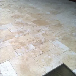 Jasne, beżowe płytki podłogowe, częściowo ułożone na zewnątrz budynku, widoczny fragment podłoża pod płytkami.