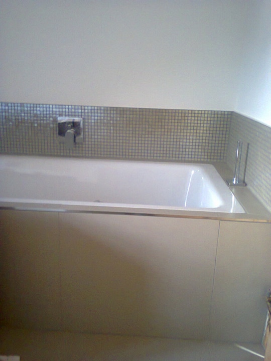 Biała wanna obudowana panelami, z mozaiką z drobnych płytek nad wanną i chromowaną baterią, widok z niskiego kąta.