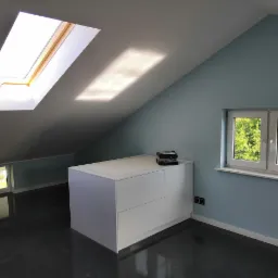 Jasne, nowocześnie wykończone poddasze ze skośnym sufitem, oknem dachowym i małym oknem w ścianie szczytowej. Na podłodze widoczny połyskujący lakier. Biała zabudowa meblowa z szufladami pod oknem.