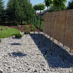 Ogród z białymi kamieniami, kulistymi drzewami przy drewnianym płocie i ozdobnymi czosnkami, oświetlony małymi lampami.