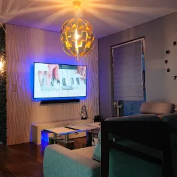 Nowoczesny salon z dekoracyjną ścianą 3D, podświetlonym telewizorem, designerską lampą i zegarem ściennym, widok na sąsiednie pomieszczenie z akwarium.