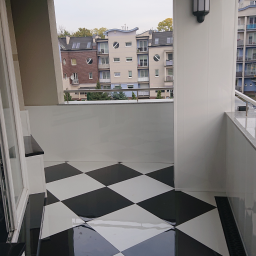 Company - Błyszcząca, czarno-biała podłoga w szachownicę na balkonie z widokiem na budynki mieszkalne, oświetlona zewnętrzną lampą ścienną.