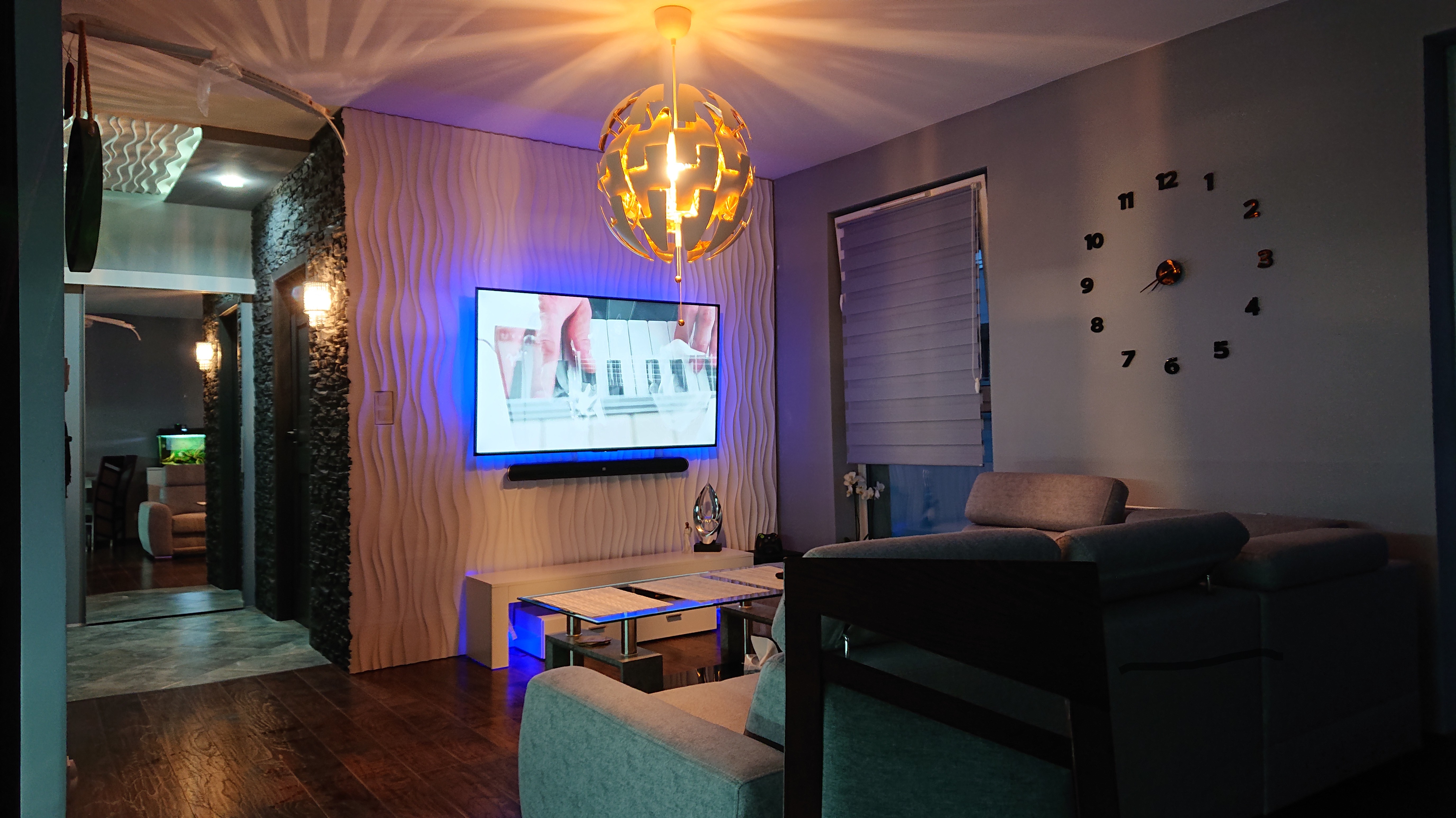 Nowoczesny salon z dekoracyjną ścianą 3D, podświetlonym telewizorem, designerską lampą i zegarem ściennym, widok na sąsiednie pomieszczenie z akwarium.